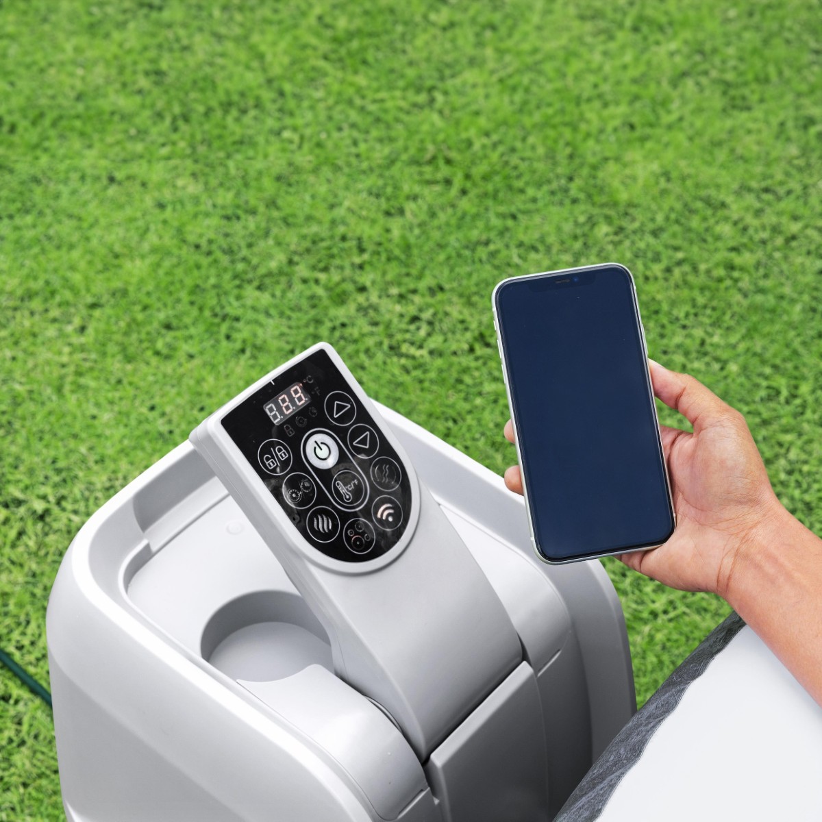 Dłoń trzymająca smartfon obok panelu sterowania pompy jacuzzi ustawionej na trawie. Panel posiada cyfrowy wyświetlacz oraz przyciski funkcyjne, w tym ikonę Wi-Fi.